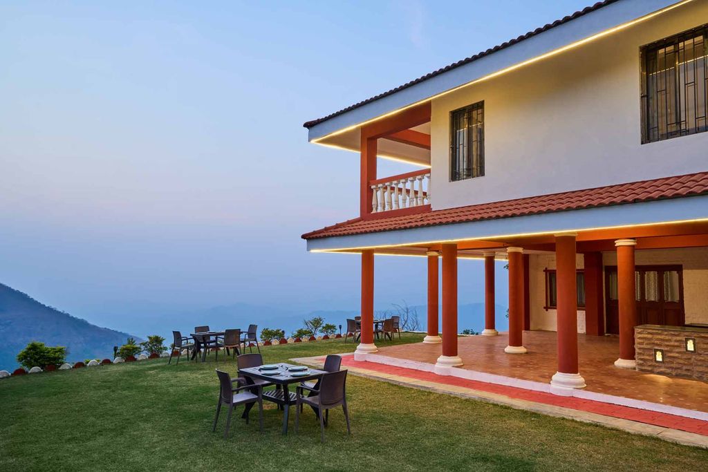 Lit veranda and lawn with dining tables at dusk, hills beyond